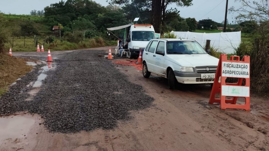 A imagem mostra uma estrada de terra com alguns trechos cobertos por cascalho. No lado direito, há um carro branco parado ao lado de um cavalete laranja com uma placa que diz “FISCALIZAÇÃO AGROPECUÁRIA”. Atrás do carro, há um furgão branco com um toldo estendido, sob o qual estão pessoas sentadas em cadeiras, aparentemente responsáveis pela fiscalização. Cones de sinalização estão espalhados pela via, delimitando o local da abordagem. O cenário é rural, com vegetação, árvores e uma pequena construção ao fundo. O dia está nublado e o chão está molhado, com poças de água.