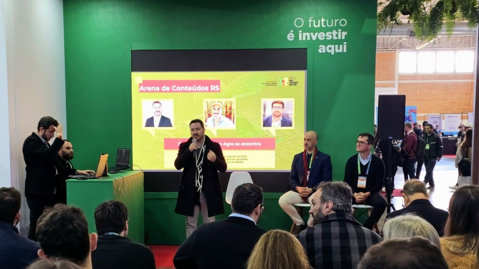 Foto de um homem em pé falando ao microfone. Outros dois painelistas estão sentados observando. Parte da plateia pode ser vista de costas, em Painel sobre Centro Agro na Gramado Summit 2025