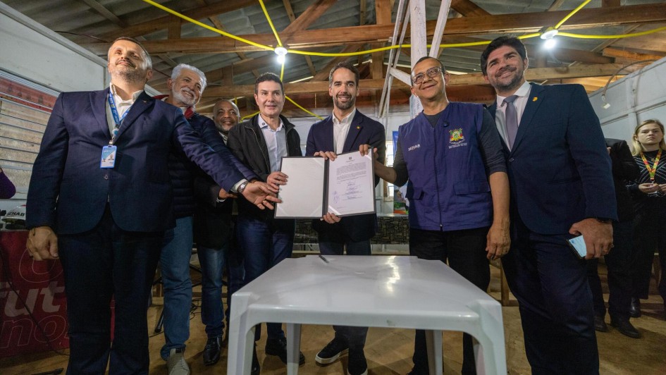 Na foto, sete homens estão em pé, sorrindo, posando para foto. Ao centro, Leite segura um documento aberto, visível para a câmera, enquanto os outros encostam as mãos no papel ou no ombro uns dos outros. Eles estão em um espaço interno simples, com teto de madeira e fios amarelos cruzando no alto. À frente deles há uma mesa branca de plástico, vazia, exceto por uma caneta. Ao fundo, outras pessoas observam a cena.