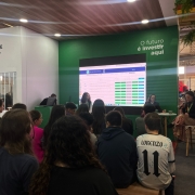 Há dezenas de crianças de costas, sentadas, assistindo uma apresentação. No palco há quatro pessoas. Dias mulheres fazem a apresentação, enquanto uma menina assiste e um homem está sentado de frente ao um notebook. Há um painel com informações atrás deles.