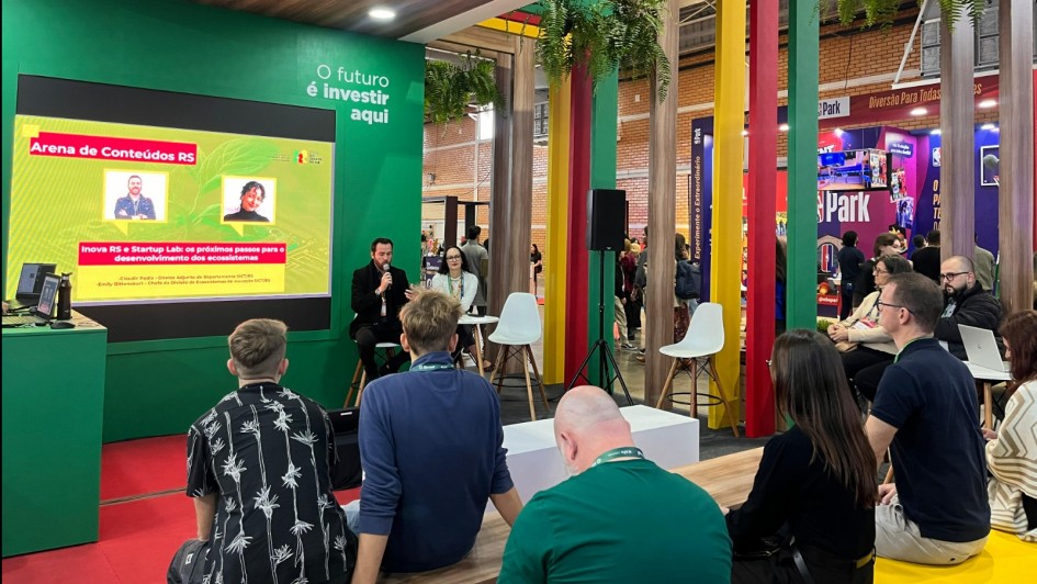 Foto mostra um evento de tecnologia e inovação em ambiente interno moderno, com destaque para uma apresentação no "Arena de Conteúdos RS" sobre "Inova RS e Startup Lab". Dois palestrantes - um homem de terno com microfone e uma mulher - apresentam para uma plateia de cerca de 20 pessoas sentadas em cadeiras brancas e amarelas, em um espaço decorado com pilares coloridos (verde, amarelo e vermelho), teto de madeira, plantas suspensas e outros estandes ao fundo, criando uma atmosfera contemporânea de networking e empreendedorismo no Rio Grande do Sul.