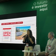 Há duas pessoas em um palco. Uma mulher segura um microfone na mão. Ela está de pé e parece falar. Um homem está sentado, e olha para ela. Ao fundo há um painel de apresentação em uma parede verde.