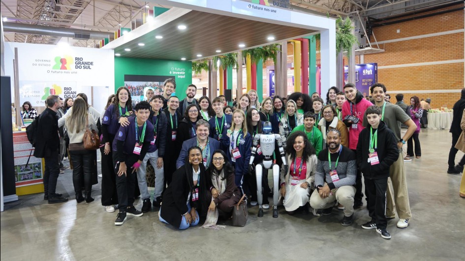 Foto de um grande grupo de pessoas reunidas em frente ao estande do Governo do Rio Grande do Sul, em um evento realizado em um pavilhão coberto. A maioria usa crachás coloridos e colares verdes. Ao centro, há um robô humanoide com braços estendidos, rodeado por pessoas sorridentes, muitas delas jovens. O ambiente tem decoração moderna, com cores vivas, plantas e letreiros.