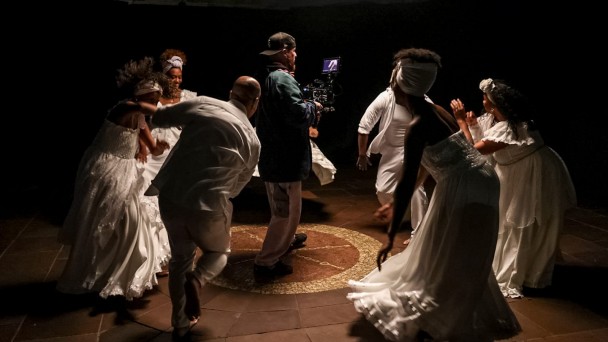 Imagem mostra grupo de atores dançando ao redor de cinegrafista. A luz está focada no meio do grupo, que usa roupas brancas