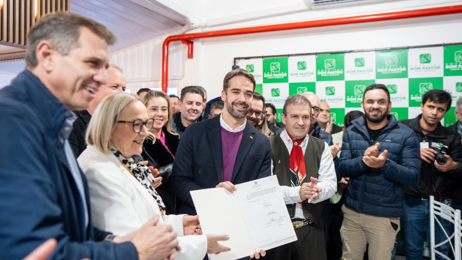 A imagem retrata um evento público em um ambiente interno, onde um grupo de pessoas está reunido, a maioria sorrindo e olhando para o centro da cena.
Em destaque no centro, um homem com barba e blazer escuro, parecendo ser uma figura de autoridade, segura um documento branco, que aparenta ser um certificado ou contrato, e sorri para a câmera. Ele está cercado por outras pessoas.
À esquerda do homem central, uma mulher de cabelos claros e óculos, vestindo um blazer branco, também sorri e parece interagir com o documento ou com o homem ao seu lado. Ao lado dela, parcialmente visível, um homem de camisa escura sorri.
À direita do homem central, um homem mais velho com um chapéu e um lenço vermelho no pescoço está aplaudindo. Atrás deles, há várias outras pessoas, a maioria homens, observando e alguns também aplaudindo.
O fundo da imagem mostra uma parede verde com logotipos repetidos, que parecem ser o nome "BOM PASTOR" e um símbolo de uma cruz, sugerindo o nome de uma instituição. Há também tubulações vermelhas visíveis e um aparelho de ar condicionado no alto da parede. A iluminação é clara e uniforme, indicando um evento bem iluminado. A atmosfera geral é de celebração.