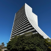 Foto de baixo pra cima do Caff, que é enquadrado de meio de lado. O prédio tem formato de rampa, com a base maior que o topo, e a cor é cinza. O céu é azul sem nuvens.