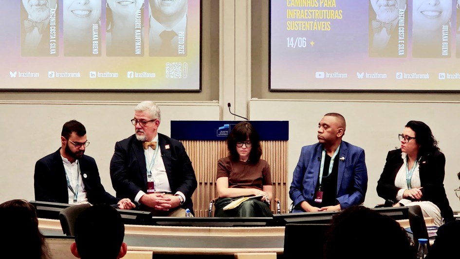 Foto de cinco pessoas estão sentadas à frente de uma plateia, participando de um painel de debate em uma sala de conferência. Atrás deles, um telão exibe os rostos e nomes dos palestrantes, com o título do evento: “Cidades resilientes e inclusivas: desafios e caminhos para infraestruturas sustentáveis”. O ambiente é formal, com todos os participantes vestidos de forma profissional.