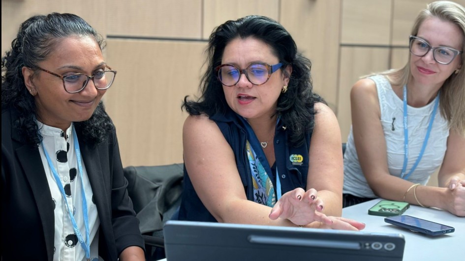 A imagem mostra três mulheres sentadas em uma mesa, aparentemente em uma reunião ou conferência.
A mulher à esquerda tem cabelo escuro e encaracolado e usa óculos. Ela está vestindo um blazer preto sobre uma blusa branca estampada e tem um crachá azul pendurado no pescoço. Ela está olhando para algo à frente com um leve sorriso.
A mulher ao centro tem cabelo escuro e usa óculos grandes e azuis. Ela está vestindo uma blusa azul-marinho e tem um crachá amarelo com a sigla "ICLEI" visível. Ela está gesticulando com a mão direita e parece estar falando ou explicando algo, olhando para uma tela de tablet à sua frente.
A mulher à direita tem cabelo loiro e usa óculos de armação clara. Ela está vestindo uma blusa branca e tem um crachá azul. Ela está olhando para a mulher do centro com atenção e um leve sorriso.
Sobre a mesa, há um tablet preto aberto, que parece ser o foco da atenção da mulher do centro. Também é possível ver um ou dois smartphones sobre a mesa. O ambiente parece ser uma sala de conferências com paredes claras.