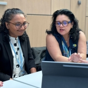 A imagem mostra três mulheres sentadas em uma mesa, aparentemente em uma reunião ou conferência.

A mulher à esquerda tem cabelo escuro e encaracolado e usa óculos. Ela está vestindo um blazer preto sobre uma blusa branca estampada e tem um crachá azul pendurado no pescoço. Ela está olhando para algo à frente com um leve sorriso.

A mulher ao centro tem cabelo escuro e usa óculos grandes e azuis. Ela está vestindo uma blusa azul-marinho e tem um crachá amarelo com a sigla "ICLEI" visível. Ela está gesticulando com a mão direita e parece estar falando ou explicando algo, olhando para uma tela de tablet à sua frente.

A mulher à direita tem cabelo loiro e usa óculos de armação clara. Ela está vestindo uma blusa branca e tem um crachá azul. Ela está olhando para a mulher do centro com atenção e um leve sorriso.

Sobre a mesa, há um tablet preto aberto, que parece ser o foco da atenção da mulher do centro. Também é possível ver um ou dois smartphones sobre a mesa. O ambiente parece ser uma sala de conferências com paredes claras.