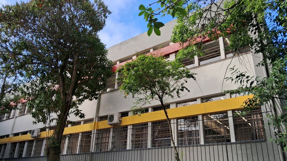 Esta é a fachada da Escola Estadual de Ensino Fundamental Antônio Vieira, localizada em Novo Hamburgo. O prédio tem dois andares, várias janelas e unidades de ar-condicionado instaladas. Ele é cercado por uma grade metálica e há vegetação ao redor. A parte superior do edifício tem uma faixa vermelha, enquanto a parte inferior exibe uma faixa amarela. O céu está azul com algumas nuvens, trazendo uma atmosfera agradável. Parece ser um ambiente escolar bem estruturado!
