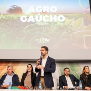A imagem mostra um evento relacionado ao agro gaúcho, realizado em 16 de outubro de 2023. No palco, há um grande telão exibindo esse título, acompanhado de uma imagem agrícola com o sol ao fundo. Abaixo da tela, seis pessoas estão sentadas em uma mesa com microfones e garrafas de água, enquanto uma pessoa está em pé, falando ao público. O ambiente sugere uma conferência ou reunião focada em discussões sobre agricultura no Rio Grande do Sul. Parece ser um evento importante para debater desafios e oportunidades do setor.