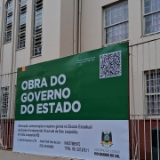 A imagem mostra um prédio com várias janelas e um cartaz verde e branco preso a uma cerca na frente do edifício. O cartaz contém texto em português, um QR code e informações sobre um projeto governamental de manutenção e reparos na Escola Estadual de Ensino Fundamental Visconde de São Leopoldo, localizada em São Leopoldo, Rio Grande do Sul, Brasil. Também há detalhes sobre a empresa responsável pela obra e o investimento total, que é de R$ 1.287.670,11. O QR code convida os espectadores a escaneá-lo para obter mais informações. A cena destaca um investimento público na infraestrutura escolar da região.
