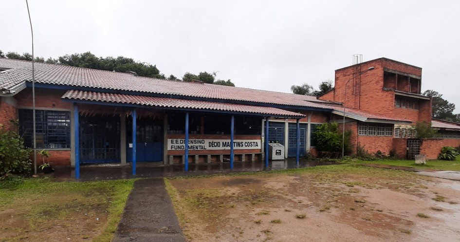 A imagem mostra a fachada da Escola Estadual de Ensino Fundamental Décio Martins Costa. O prédio tem paredes de tijolos vermelhos e um telhado de telhas. Na frente da escola, há uma placa azul e branca com o nome da instituição. As janelas possuem grades metálicas, e as portas são azuis. No lado direito da imagem, há uma lixeira preta e branca. O chão parece estar molhado, indicando que choveu recentemente. A cena transmite um ambiente escolar organizado e seguro.
