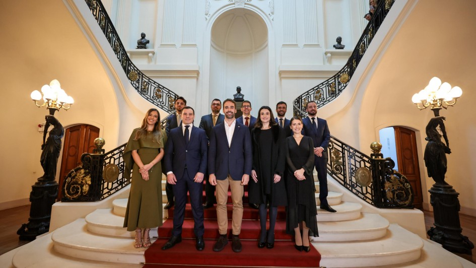 A imagem mostra um grupo de pessoas em uma escadaria grandiosa dentro de um edifício elegante. A arquitetura apresenta detalhes ornamentados, esculturas e luminárias decorativas. Os presentes estão vestidos formalmente, indicando um evento especial ou cerimônia oficial.