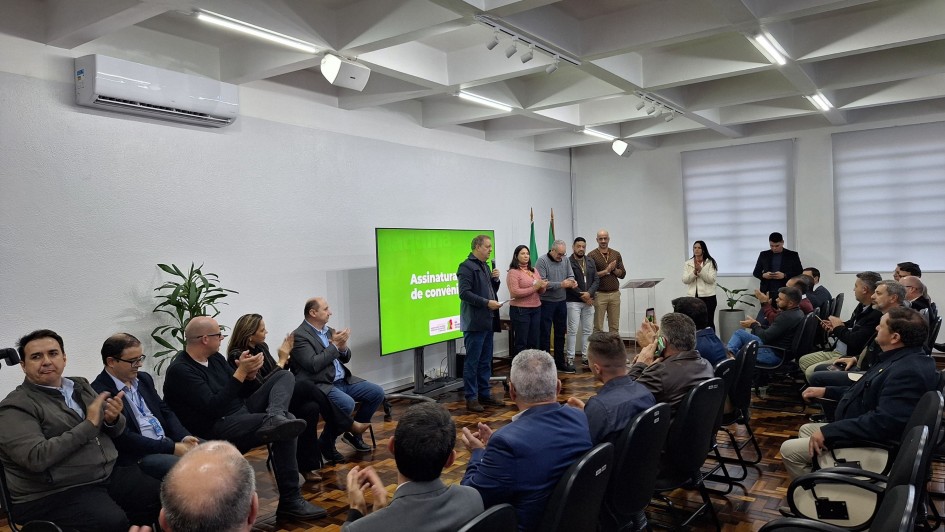 A imagem mostra um grupo de pessoas em um ambiente interno, que parece ser uma sala de reuniões ou um auditório pequeno. Há um palco ou área de apresentação na frente, onde algumas pessoas estão de pé, e uma tela grande exibe o texto "Assinatura do convênio". A maioria das pessoas está sentada em cadeiras dispostas em fileiras, de frente para a área de apresentação. Muitas delas estão aplaudindo. A sala tem paredes claras, iluminação de teto e ar condicionado. No canto esquerdo da imagem, uma planta verde contribui para a decoração. O ambiente geral sugere um evento formal, possivelmente uma cerimônia de assinatura ou anúncio importante.