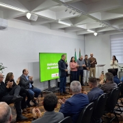 A imagem mostra um grupo de pessoas em um ambiente interno, que parece ser uma sala de reuniões ou um auditório pequeno. Há um palco ou área de apresentação na frente, onde algumas pessoas estão de pé, e uma tela grande exibe o texto "Assinatura do convênio". A maioria das pessoas está sentada em cadeiras dispostas em fileiras, de frente para a área de apresentação. Muitas delas estão aplaudindo. A sala tem paredes claras, iluminação de teto e ar condicionado. No canto esquerdo da imagem, uma planta verde contribui para a decoração. O ambiente geral sugere um evento formal, possivelmente uma cerimônia de assinatura ou anúncio importante.