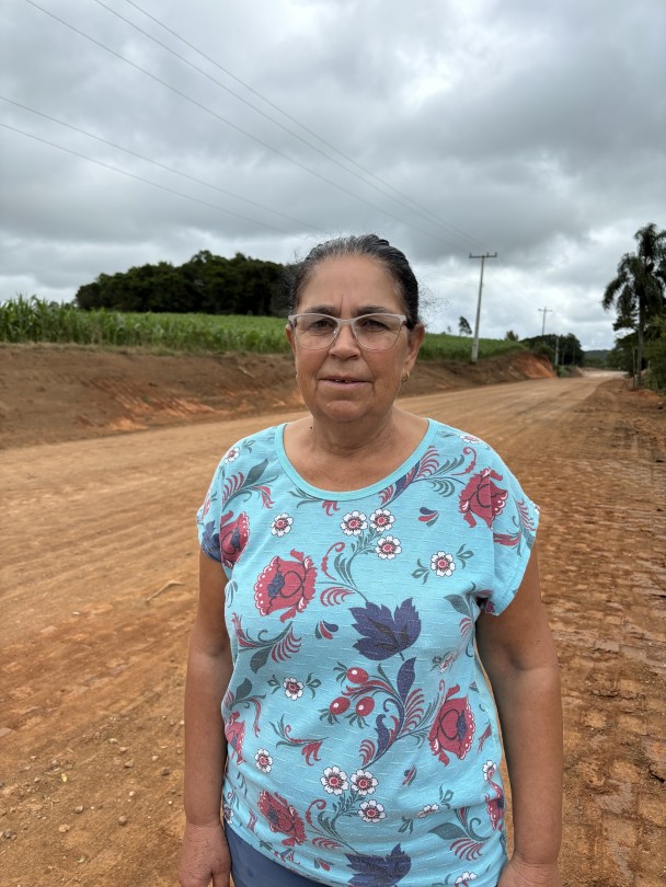 A imagem apresenta uma mulher de meia-idade, com pele clara e cabelos escuros parcialmente presos, usando óculos de armação transparente. Ela veste uma camiseta de manga curta azul-clara com estampas florais vibrantes em tons de vermelho, branco e azul-escuro. A mulher está em pé, posicionada do peito para cima, olhando diretamente para a câmera com uma expressão neutra.

Ela se encontra em um ambiente externo, em uma estrada de terra batida de cor avermelhada, que se estende ao fundo. No lado esquerdo da imagem, é possível ver um campo de cultivo verde, provavelmente milho, e algumas árvores distantes. No lado direito, há mais vegetação, um poste de energia elétrica e fios que cruzam o céu nublado. A iluminação é difusa, indicando um dia nublado.