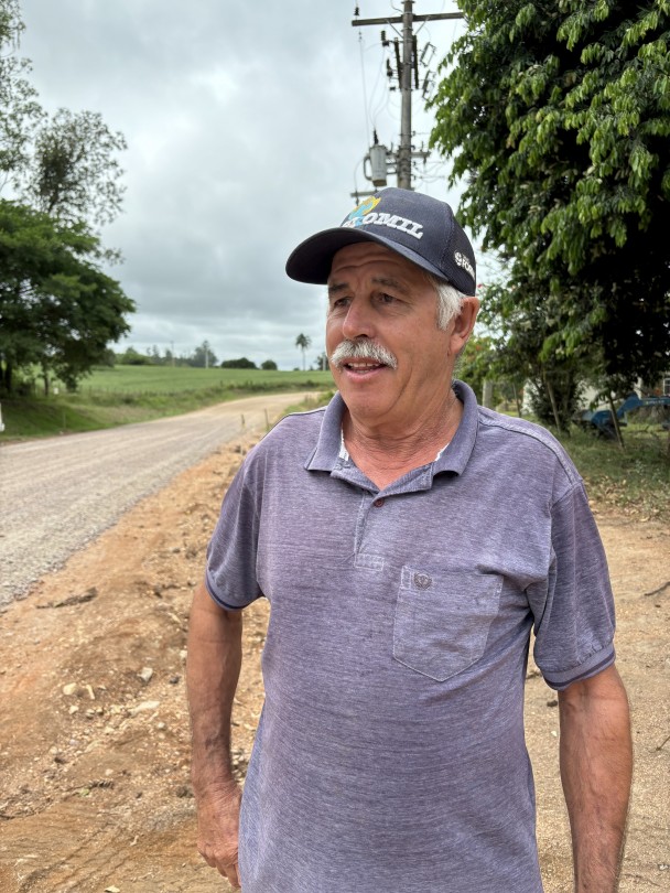 A imagem mostra um homem de meia-idade, com pele clara, cabelos grisalhos visíveis nas laterais e um bigode branco. Ele está usando um boné escuro com a aba virada para trás, que tem a palavra "COMIL" parcialmente visível em cores vibrantes, e uma camiseta polo de cor azul acinzentada com um bolso no peito. Ele está posicionado em um ângulo de três quartos, olhando para o lado direito da imagem com uma expressão pensativa.

O cenário atrás dele é uma estrada de terra batida de cor avermelhada. No lado esquerdo da estrada, há um campo verde com algumas árvores ao fundo. No lado direito, próximo ao homem, há um poste de energia elétrica com vários fios e um emaranhado de cabos, e algumas árvores com folhagem densa. O céu está nublado, indicando um dia com pouca luz solar direta.