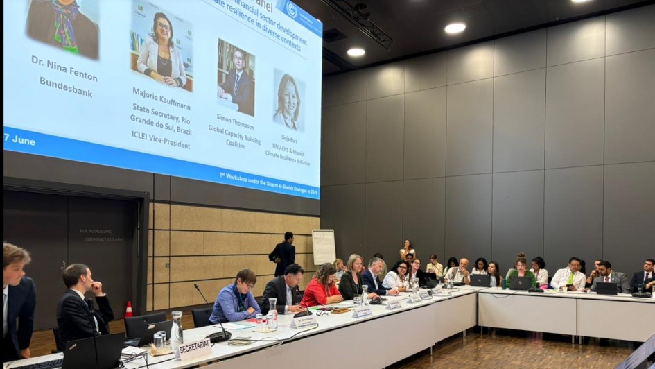 A imagem mostra uma conferência com participantes reunidos ao redor de uma mesa em formato de U. Há placas de identificação, laptops e microfones na mesa, e um grande telão ao fundo exibe informações sobre um painel de discussão relacionado ao financiamento climático.