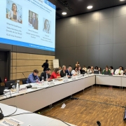 A imagem mostra uma conferência com participantes reunidos ao redor de uma mesa em formato de U. Há placas de identificação, laptops e microfones na mesa, e um grande telão ao fundo exibe informações sobre um painel de discussão relacionado ao financiamento climático.