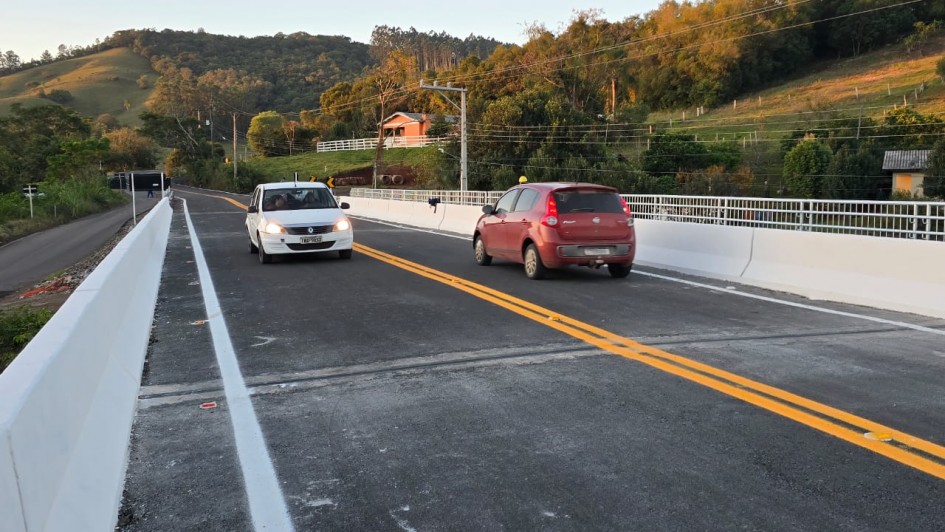 A imagem mostra uma ponte com dois carros trafegando em sentidos opostos. A estrutura possui barreiras brancas nas laterais e marcação central amarela. Ao fundo, há uma paisagem rural com colinas, árvores e algumas casas.
