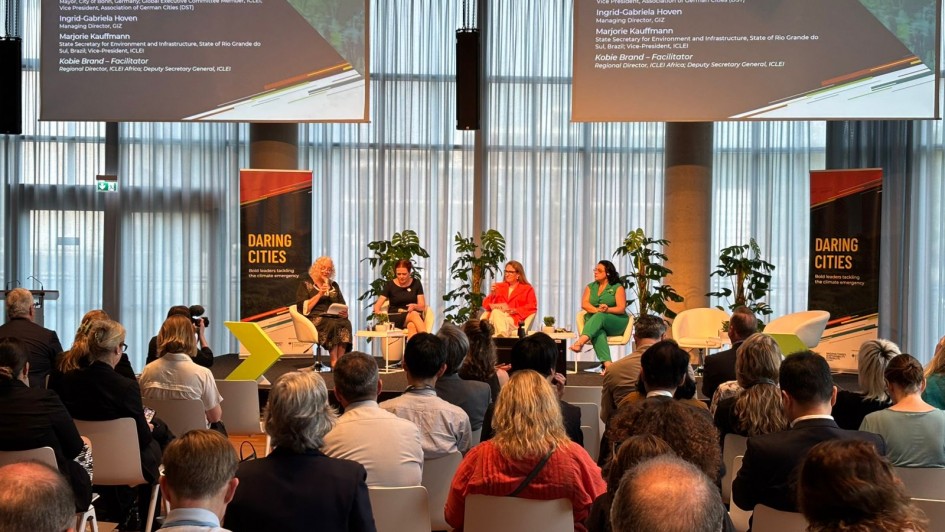 Foto de uma conferência, tirada do fundo de um auditório fechado. No primeiro plano, está o público sentado de costas. Ao fundo, quatro mulheres sentadas em cadeiras no palco, com banners ao lado.