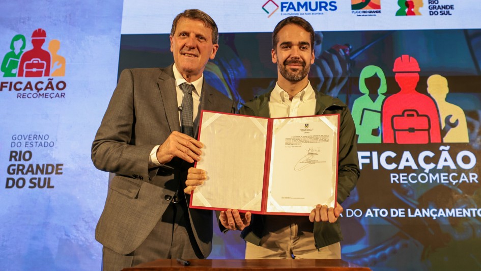 Gilmar e Leite posam para foto, de frente, sorrindo para a câmera. Ambos estão de pé e seguram um documento assinado.
