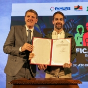 Gilmar e Leite posam para foto, de frente, sorrindo para a câmera. Ambos estão de pé e seguram um documento assinado.