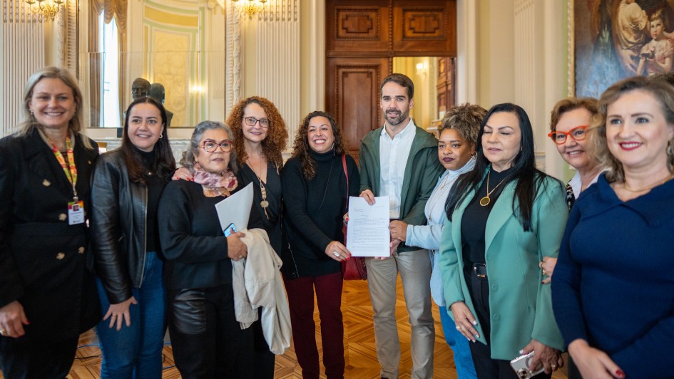 A imagem mostra um grupo de onze pessoas, a maioria mulheres, em um ambiente interno que parece ser um prédio governamental ou histórico.
No centro da imagem, um homem com barba, vestindo camisa social branca e blazer verde-oliva, está de pé e segurando um documento. Ele está ladeado por mulheres de diferentes etnias e idades.
À esquerda do homem, há cinco mulheres. A primeira, da esquerda para a direita, usa uma blusa preta e um colar de identificação. A segunda veste uma jaqueta preta. A terceira, com óculos e cabelo grisalho, segura um documento. A quarta tem cabelo cacheado e usa óculos. A quinta está com um casaco preto.
À direita do homem, há cinco mulheres. A primeira (imediatamente à direita dele) tem cabelo crespo. A segunda usa um blazer verde-água e um colar dourado. A terceira tem óculos e cabelo ruivo. A quarta mulher, no canto direito da imagem, tem cabelo claro e usa uma blusa azul-marinho.
O ambiente ao fundo é ornamentado, com tetos altos, portas de madeira escuras e quadros nas paredes. Há também um espelho grande refletindo parte do ambiente.