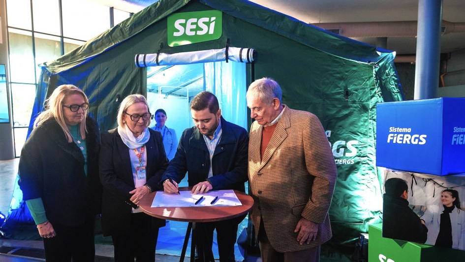 A imagem mostra quatro pessoas em pé ao redor de uma pequena mesa redonda, com uma pessoa assinando um documento. Eles estão em frente a uma tenda verde com o logotipo "SESI". Também há caixas azuis com o texto "Sistema Fiergs" visíveis ao fundo.
