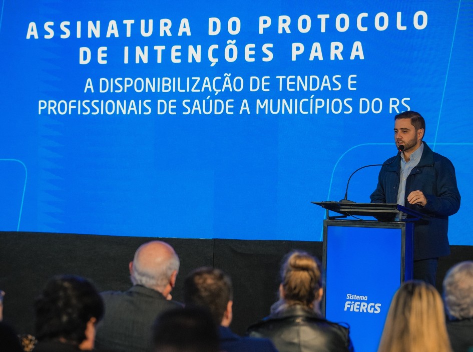 A imagem mostra Gabriel Souza em um púlpito com o texto "ASSINATURA DO PROTOCOLO DE INTENÇÕES PARA A DISPONIBILIZAÇÃO DE TENDAS E PROFISSIONAIS DE SAÚDE A MUNICÍPIOS DO RS" exibido em uma tela grande atrás dele. O púlpito tem o logotipo "Sistema FIERGS". Há pessoas sentadas, voltadas para o palco.