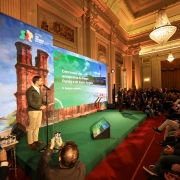 A imagem retrata um evento formal em um grande salão, onde um homem está discursando em um palco e uma plateia o assiste.

No centro da imagem, em um palco elevado coberto por um carpete verde, um homem de barba, vestindo uma jaqueta verde-oliva, camisa branca e calças cáqui, está de pé em frente a um microfone. Ele parece estar apresentando algo, talvez utilizando um controle remoto na mão direita.

Atrás dele, há um grande telão dividido em duas seções. A seção da esquerda exibe uma imagem de ruínas históricas e, no canto superior esquerdo, um logotipo com "GOVERNO RIO GRANDE DO SUL". A seção da direita mostra um texto que diz "Concessão dos aeroportos de Passo Fundo e de Santo Ângelo" e abaixo "Projeto revisado".

À direita do palco, há uma grande plateia sentada em cadeiras, com muitas pessoas observando o orador. Algumas pessoas, no lado esquerdo da imagem, estão em pé e tirando fotos com seus celulares.

O salão é grandioso, com teto alto, paredes ornamentadas, colunas e lustres de cristal suspensos, sugerindo um ambiente histórico ou governamental. O piso à frente do palco é vermelho e há iluminação focada no palco.