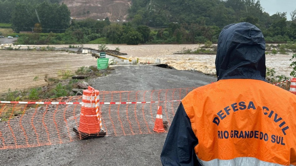 Imagem mostra homem da Defesa Civil do Rio Grande do Sul olhando para água de rio que cobre ponte.