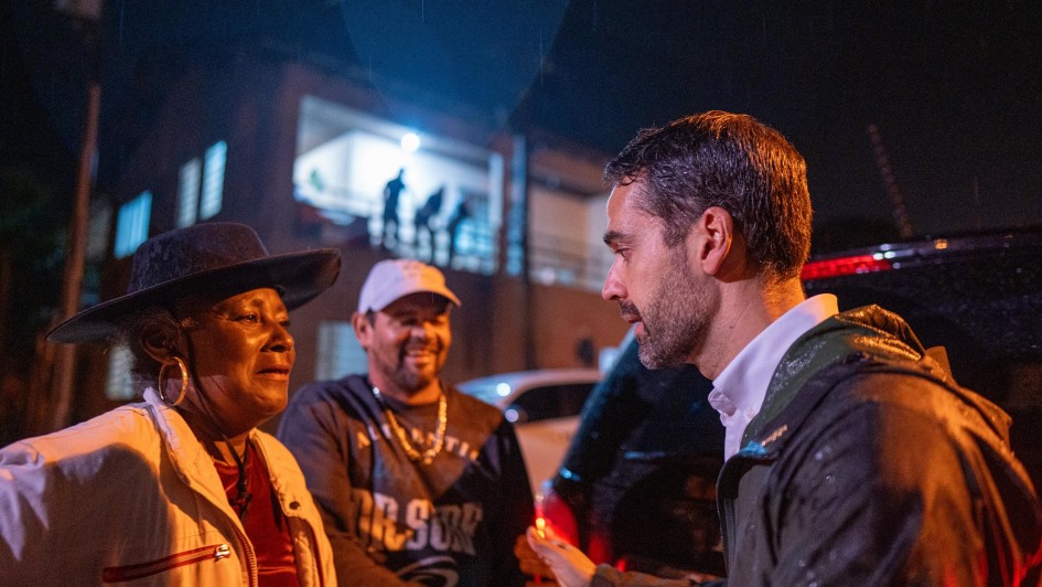 A imagem mostra uma cena noturna, sob chuva fina, com um homem de barba (o Governador) conversando com uma mulher mais velha e um homem ao fundo.
O homem da direita, com barba, veste uma camisa social branca e um casaco escuro, que parece molhado pela chuva. Ele está olhando e falando com a mulher à sua frente, e tem uma pequena chama (talvez de um isqueiro ou vela) perto de sua mão direita.
A mulher à esquerda, mais velha, usa um chapéu escuro, brincos de argola e uma jaqueta clara sobre uma blusa vermelha. Ela parece emocionada, com a expressão facial e os olhos marejados, olhando para o homem da direita.
Atrás da mulher, um homem de boné claro e camiseta escura com uma estampa sorri e observa a interação.
O fundo é escuro, com silhuetas de pessoas em uma varanda iluminada e parte de um veículo escuro visível à direita. Pequenos pontos brilhantes na imagem sugerem a chuva caindo.