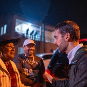 A imagem mostra uma cena noturna, sob chuva fina, com um homem de barba (o Governador) conversando com uma mulher mais velha e um homem ao fundo.

O homem da direita, com barba, veste uma camisa social branca e um casaco escuro, que parece molhado pela chuva. Ele está olhando e falando com a mulher à sua frente, e tem uma pequena chama (talvez de um isqueiro ou vela) perto de sua mão direita.

A mulher à esquerda, mais velha, usa um chapéu escuro, brincos de argola e uma jaqueta clara sobre uma blusa vermelha. Ela parece emocionada, com a expressão facial e os olhos marejados, olhando para o homem da direita.

Atrás da mulher, um homem de boné claro e camiseta escura com uma estampa sorri e observa a interação.

O fundo é escuro, com silhuetas de pessoas em uma varanda iluminada e parte de um veículo escuro visível à direita. Pequenos pontos brilhantes na imagem sugerem a chuva caindo.