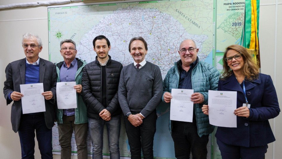 Na imagem aparecem seis pessoas sorridentes, cinco homens e uma mulher. Eles posam para uma foto em frente a um grande mapa rodoviário. As duas pessoas do lado esquerdo da foto e as duas que estão do lado direito seguram papéis. A mulher à direita usa óculos e um casaco azul-escuro.