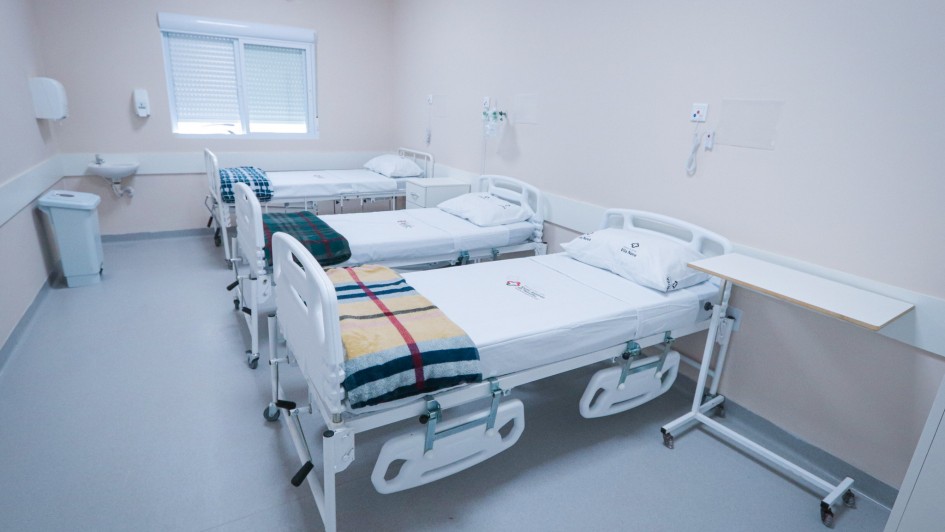 Imagem mostra um quarto de hospital claro e moderno com três camas brancas arrumadas. Há um ar condicionado na parede superior e uma janela com persianas.