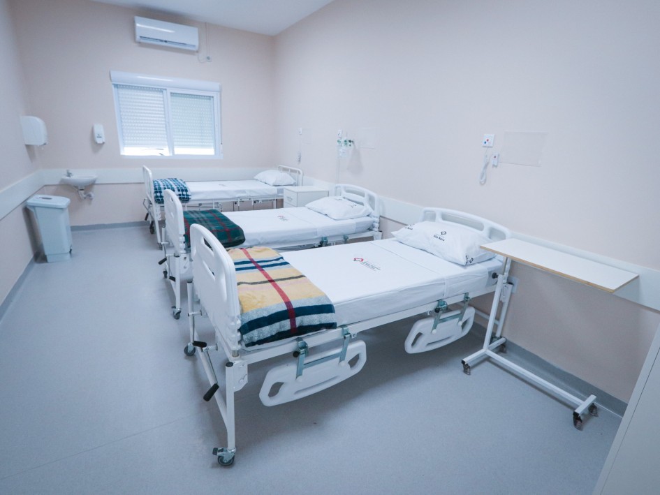Imagem mostra um quarto de hospital claro e moderno com três camas brancas arrumadas. Há um ar condicionado na parede superior e uma janela com persianas.