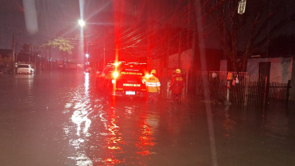 Uma rua noturna está completamente alagada, com a água refletindo as luzes. Um veículo de emergência com luzes vermelhas intermitentes está parado na água, e dois bombeiros estão em pé na rua inundada, aparentemente avaliando a situação. A imagem transmite a intensidade do alagamento e a presença das forças de segurança.