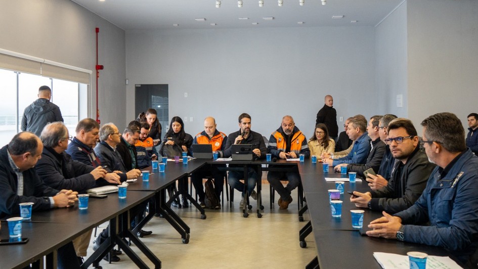 A imagem mostra uma reunião formal com várias pessoas sentadas ao redor de uma mesa, algumas usando jaquetas laranja, possivelmente representando uma equipe ou órgão específico.