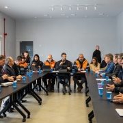 A imagem mostra uma reunião formal com várias pessoas sentadas ao redor de uma mesa, algumas usando jaquetas laranja, possivelmente representando uma equipe ou órgão específico.