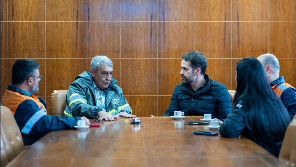 Foto de cinco pessoas sentadas ao redor de uma mesa de madeira em uma sala de reuniões com painéis de madeira ao fundo. Sobre a mesa, há xícaras de café, pires, celulares e pequenos objetos.
