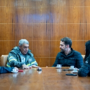 Foto de cinco pessoas sentadas ao redor de uma mesa de madeira em uma sala de reuniões com painéis de madeira ao fundo. Sobre a mesa, há xícaras de café, pires, celulares e pequenos objetos.