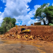 A imagem mostra uma escavadeira amarela operando em um terreno com pedras e terra, em meio a uma área com vegetação e construções ao fundo. A cena representa trabalhos de desassoreamento realizados no município de Barra do Rio Azul, voltados à prevenção de enchentes.