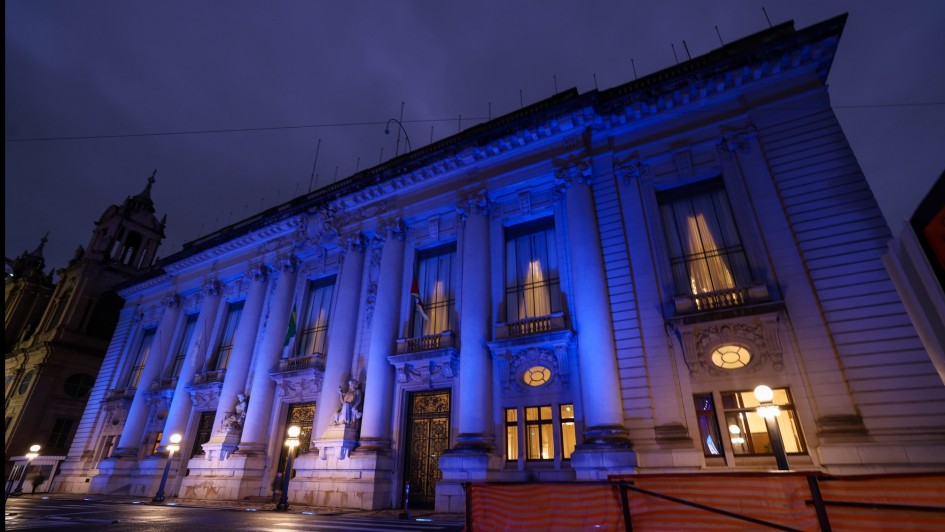 A imagem mostra o Palácio Piratini, sede do governo do Rio Grande do Sul, iluminado com luz azul durante a noite. A fachada neoclássica com colunas e esculturas está em destaque. A iluminação azul faz referência ao Dia Mundial do Refugiado.