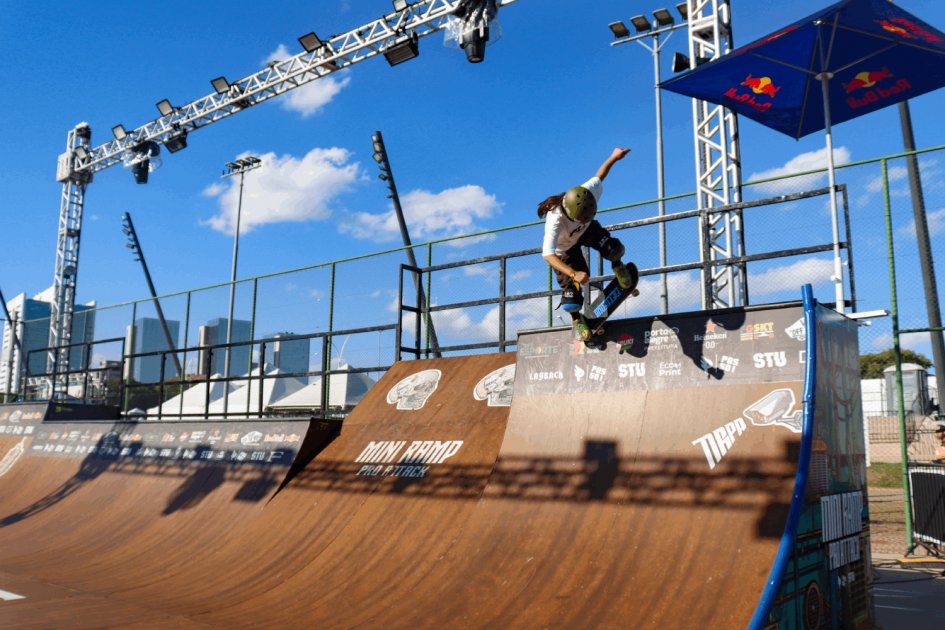 Imagem mostra skatista fazendo manobra em rampa. Ao fundo, no alto, céu azul e algumas nuvens, e abaixo aparecem prédios. A pista de skate tem refletores no alto.