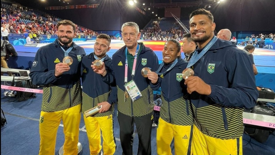Imagem mostra cinco atletas com abrigo de calça amarela e blusa azul, com listras amarelas. Todos exibem felizes suas medalhas. Ao fundo, tatames de judô com pessoas lutando e ao fundo o público.