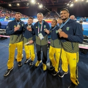 Imagem mostra cinco atletas com abrigo de calça amarela e blusa azul, com listras amarelas. Todos exibem felizes suas medalhas. Ao fundo, tatames de judô com pessoas lutando e ao fundo o público.