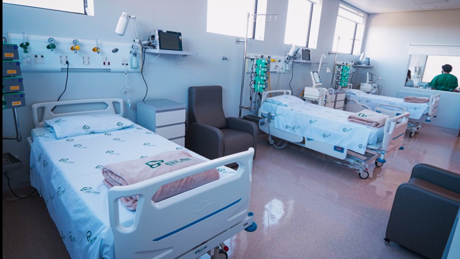 A imagem retrata um quarto de hospital com três leitos de UTI totalmente equipados. Cada cama possui roupa de cama com a marca do SUS e do Governo Federal, e está acompanhada de equipamentos médicos como monitores, bombas de infusão e suportes de soro. O ambiente é limpo, organizado e pronto para atender pacientes em cuidados intensivos.