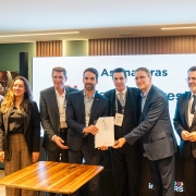 A imagem mostra um grupo de oito pessoas reunidas em um ambiente formal, com algumas segurando um documento. Ao fundo, há uma grande tela exibindo parcialmente os textos "Assinaturas" e "RS". Há também uma mesa de madeira em primeiro plano.
